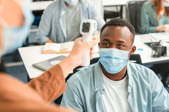 Prevention And Precautions. Woman using digital medical non-contact infrared electronic thermometer gun, measuring and screening body temperature of African American student wearing surgical face mask