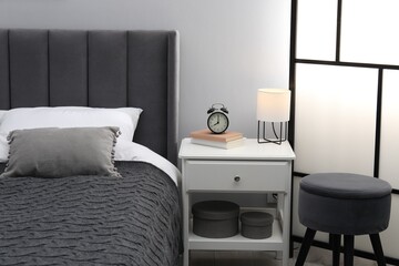 Bedside table with lamp, alarm clock and books near bed indoors