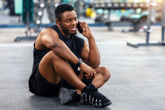 Smiling african american athletic man resting after workout in gym, copy space. Muscular black guy sitting on floor in gym, holding bottle with protein or water and talking on mobile phone