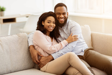 Obraz premium Happy Loving Family. Beautiful african american man and woman cuddling and relaxing on the couch at home in living room, smiling couple looking at camera and posing, spending leisure time together