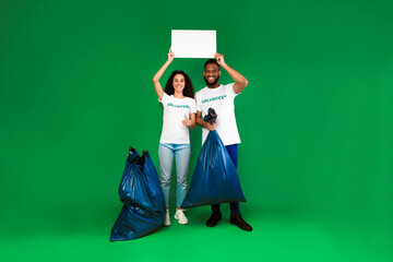 Two Diverse Volunteers Holding Blank Paper Board Posing With Plastic Bags Full Of Wasted Junk Standing Over Green Studio Background. Volunteering Work Concept. Mockup
