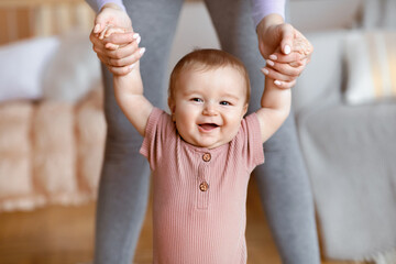 Happy little baby boy taking first steps with mother help, home interior, close up. Laughing cute toddler holding mom hands, adorable blonde kid daughter walking by living room