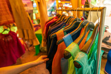 Woman shopping for colorful traditional clothes at rural Thai street market where local vendors sell handmade garments to tourists during cultural travel experience in countryside Thailand