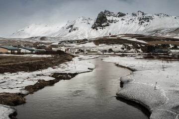 A winter landscape featuring a river flowing through a valley with snow covered mountains behind it