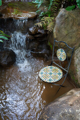 Decorative chair with colorful mosaic tiles sits in serene garden setting by small waterfall