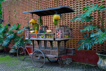 Rustic fruit cart with bananas, pineapples, and apples against brick wall, surrounded by lush green plants