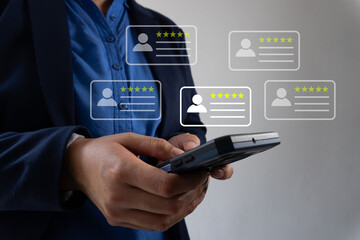 Close-up of a person's hand using a smartphone to write a positive review, with five-star icons and customer profile icons floating above the phone screen, on a grey background. 