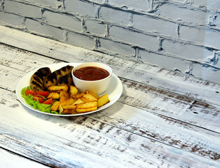 Grilled sausages, potatoes and cherry tomatoes on a lettuce leaf, next to a cup of sauce on a light wooden table.