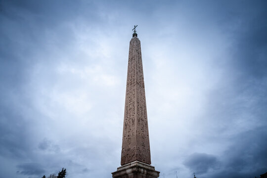 Flaminio Obelisk stands by a cloudy Roman sky in Piazza del Popolo, its granite shaft engraved with Egyptian hieroglyphs topped by a papal cross: a symbol of Rome's ancient Egyptian monuments.