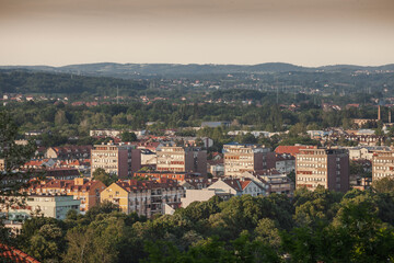 Obraz premium Elevated late-afternoon panorama of central Valjevo, Serbia, reveals dense rows of socialist-era apartment blocks and newer houses framed by rolling forested hills under a hazy summer sky.