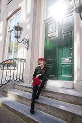 Fashionable woman standing on stairs in front of elegant green doors on streets of Amsterdam. Fashion model is holding roses in the hand and happy smiling at camera. Fashion travel concept