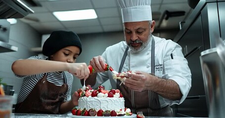 A chef and a child decorate a strawberry cake together in a professional kitchen. Warm lighting illuminates the scene - Powered by Adobe
