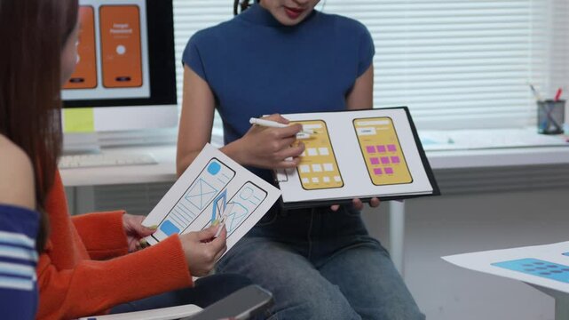 Web designers collaborating on mobile app layouts, comparing paper drafts with digital mockups on tablet during creative project planning meeting
