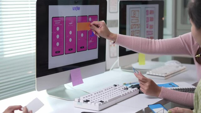 Two Asian women, probably user experience and user interface designers, are discussing and testing a new mobile app interface using sticky notes on a computer screen in an office