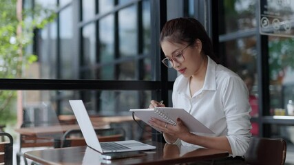 Professional Asian businesswoman concentrating on work, writing notes while using laptop at cafe table during daytime, capturing focused career moment with modern urban backdrop - Powered by Adobe