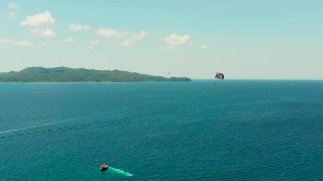 People flying on a colorful parachute towed by a motor boat, aerial view. Parasailing in blue sky.