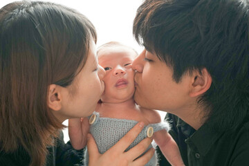 Newborn photo on a white background of a 2-week-old baby in a blue romper, gently held by a young Japanese couple. Both kiss the baby from each side. Heartwarming and professional composition.