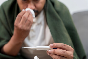 Close up of sick man holding thermometer and tissue, blowing nose and checking temperature while feeling unwell