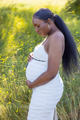 In a serene summer scene, a joyful pregnant woman embraces her baby bump outdoors, surrounded by greenery and flowers, radiating warmth, happiness, and anticipation of new beginnings