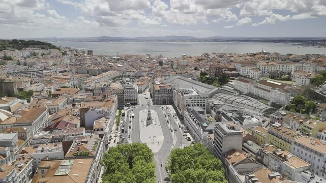 Drone flies south over Pra&ccedil;a dos Restauradores and Pra&ccedil;a do Rossio toward the sea on sunny day in Lisbon, Portugal