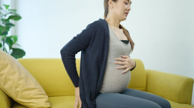 Pregnant woman in casual outfit sitting on yellow couch feeling exhausted and fatigued, holding her belly and resting her head in discomfort during the third trimester of pregnancy.