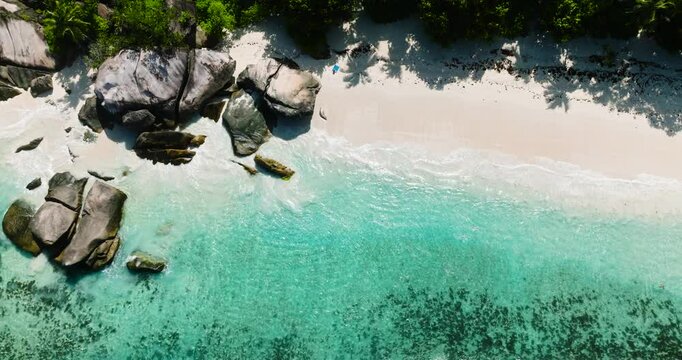 Large rock formations by the turquoise water of a tropical beach, surrounded by green foliage. Seychelles, Mahe. Roche Copra.