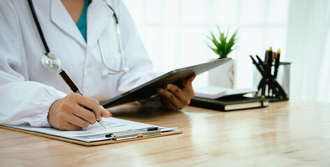 A professional female doctor, wearing a lab coat and stethoscope, sits at her desk with a tablet and clipboard, looking directly at the camera.