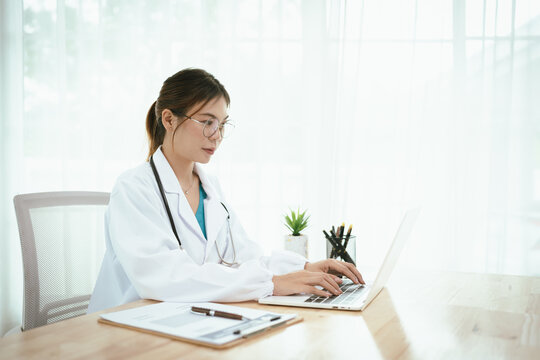 A professional female doctor, wearing a lab coat and stethoscope, sits at her desk with a tablet and clipboard, looking directly at the camera.