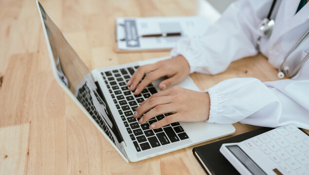 A professional female doctor, wearing a lab coat and stethoscope, sits at her desk with a tablet and clipboard, looking directly at the camera.