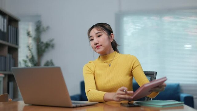 Productive young Asian businesswoman working remotely, typing on laptop while taking detailed notes in comfortable home office environment, reflecting modern professional lifestyle