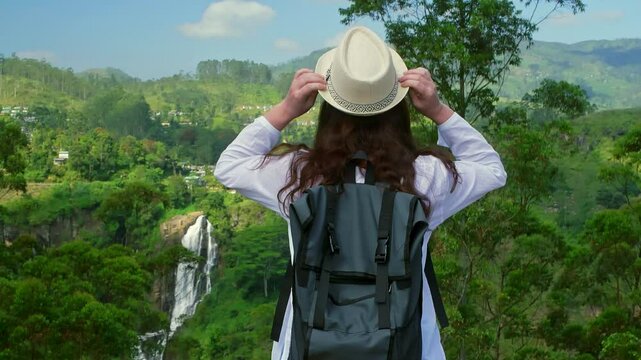 Female tourist with backpack holding her hat while admiring beautiful landscape with Ramboda Falls, surrounded by lush green tea plantations and tropical vegetation in Sri Lanka