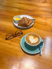 Heart-shaped latte art tops a cappuccino served with a buttery croissant and honey, set on a warm wooden table beside a pair of eyeglasses.