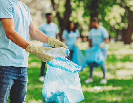 Volunteer with yellow gloves picks up plastic bottle in park, placing it in trash bag. Group of eco-friendly people blurred in background. Ideal for cleanup, sustainability, teamwork, and green action