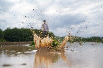A splash of muddy water rises high as a person stands blurred in the background under a cloudy sky in a natural outdoor setting.