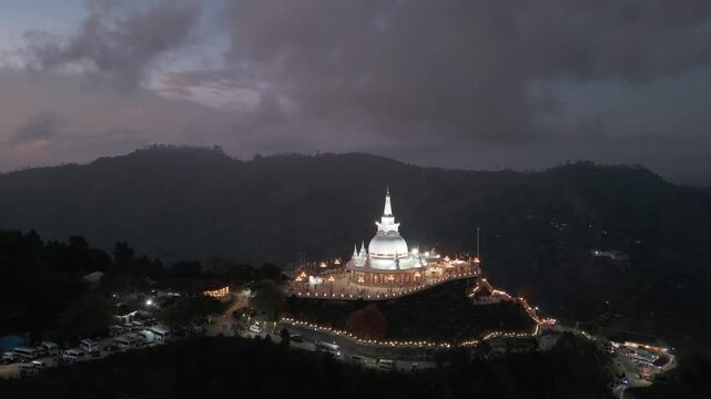 Hill top religious stupa decoratively lit up during blue hour on a mountain top in Kumbalwela, central Sri Lanka. Drone dynamic aerial.