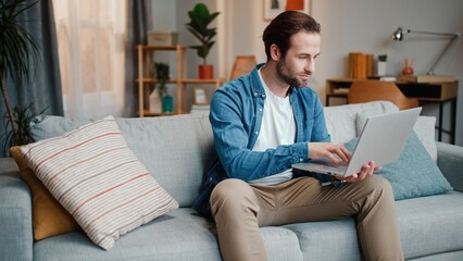 Bearded man working with laptop sitting on sofa. Male sitting on couch and working distantly. Man...