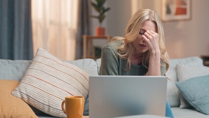 Fototapeta premium Sad woman sitting at couch and using laptop. Female sitting on sofa and drinking coffee or tea. Disappointed woman sitting in front of laptop. Female failing for work application.