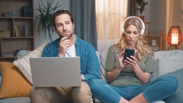 Spouses sitting on couch. Male using laptop. Young couple spending time together. Woman wearing headphones and listening music. Wife attracting attention of husband. Man and woman having fun at home.