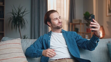 Young happy man making selfies using smartphone sitting on sofa. Cute cheerful male taking photos...