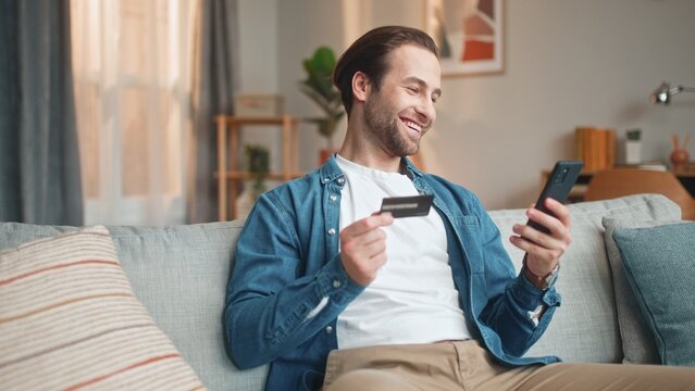 Handsome happy man holding credit card, smartphone and smiling. Young beautiful delightful male sitting on sofa buying online by phone and laughing, happy. Shopping online concept.
