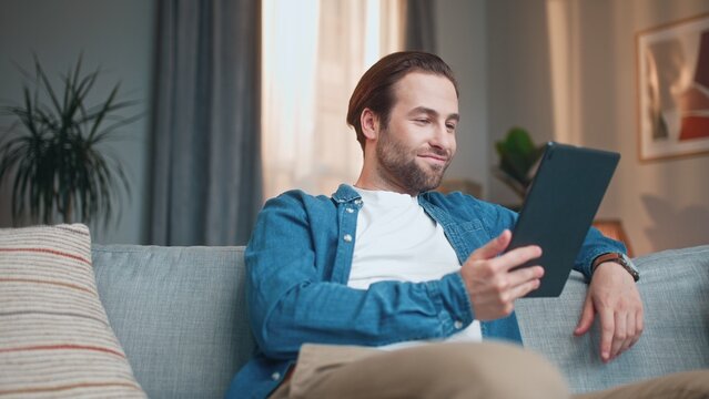 Busy charming man reading ebook using tablet online. Young beautiful male sitting on sofa in living room and reading interesting book smiling thinking. Stylish smart guy develops. Reading concept