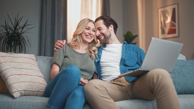 Lovely couple of beautiful man and charming woman sitting on sofa and watching film. Handsome male and cute female holding laptop talking and smiling, looking at gadget online. Entertainment concept,