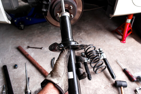  Car Shock Absorber or Strut, A mechanic hand holds a new black shock absorber or strut for replacement, surrounded by car parts and tools in a auto repair garage.