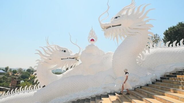 Woman tourist wear stylish white dress sightseeing explores iconic twin white dragons on temple stairs at Wat Huay Pla Kang in Chiang Rai, Thailand under clear blue sky. Famous travel phot spot