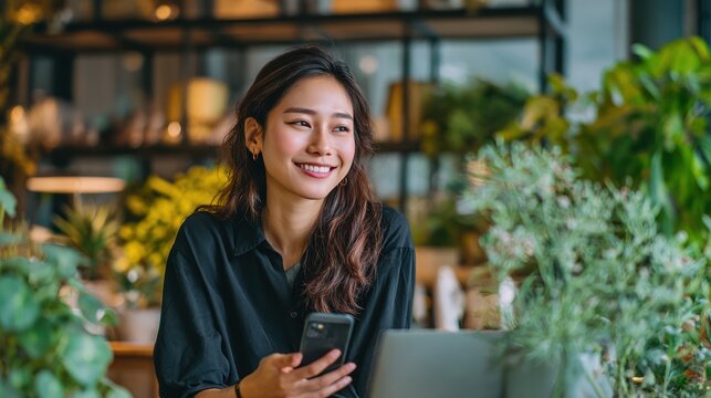 Candid image of smiling Asian teen businesswoman using mobile while seated at laptop, fresh professional look, background with office decor and plants