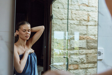 Woman wrapped in a blue towel standing by a bathroom mirror with natural stone wall background, wet hair, and relaxed expression after shower.