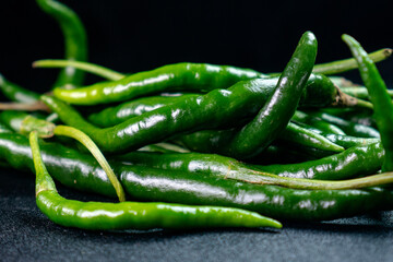 Close-up view of a pile of whole, shiny, long green chilies on a black surface and background, ideal for culinary purposes, spicy recipes, and organic vegetable promotions.