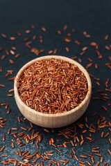 Closeup of red rice in wooden bowl with scattered grains on dark background