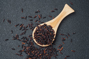 Wooden spoon filled with black rice and spilled grains on dark background