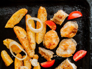 Oven pan with sliced baked potatoes, fish steaks and vegetables with onion on the table. Homemade...